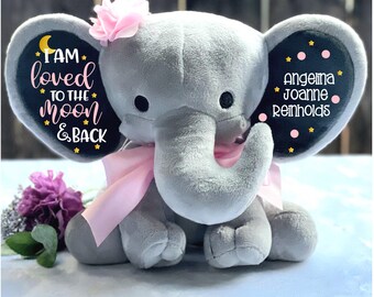 May include: A gray plush elephant toy with pink ears and a pink bow. The elephant has a black patch on each ear with the text "I am loved to the moon & back" on one ear and "Angelina Joanne Reinholds" on the other ear.