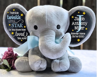 May include: A grey plush elephant toy with a light blue ribbon tied around its neck. The ears feature black fabric panels with yellow stars and white text reading Twinkle twinkle LITTLE STAR Do You Know How loved You Are? and Jacob Anthony Miller.