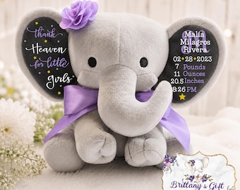 May include: A plush gray elephant toy featuring a purple satin ribbon around its neck and a purple fabric flower on its head. The ears display printed text: "thank heaven for little girls" and "Malia Milagros Rivera 02*28*2023 7 Pounds 11 Ounces 20.5 Inches 8:26 PM".