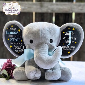 May include: A gray plush elephant toy with a light blue ribbon tied around its neck. The left ear features the text Twinkle twinkle LITTLE STAR Do You Know How loved You Are? and the right ear displays the name Jacob Anthony Miller.