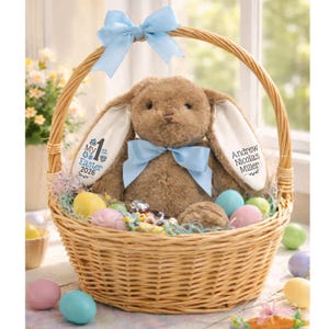 May include: A woven wicker Easter basket filled with a plush brown bunny wearing a blue bow. The basket contains colorful Easter eggs and candies. The bunny's ear has the text "My 1st Easter 2026" and the name "Andrew Nicolas Miller". A blue bow adorns the basket handle.