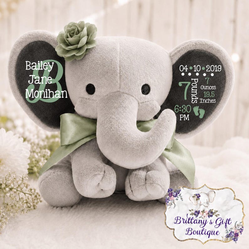 May include: A gray plush elephant toy with a sage green ribbon and a matching fabric flower on its head. The ears feature printed text including the name Bailey Jane Monihan, the date 04-10-2019, 7 pounds 7 ounces, 19.5 inches, and 6:30 PM.