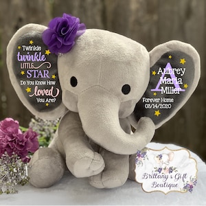 May include: A gray plush elephant toy with a purple fabric flower on its head. The left ear features the text Twinkle twinkle LITTLE STAR Do You Know How loved You Are? with yellow stars. The right ear displays the name Aubrey Maria Miller.