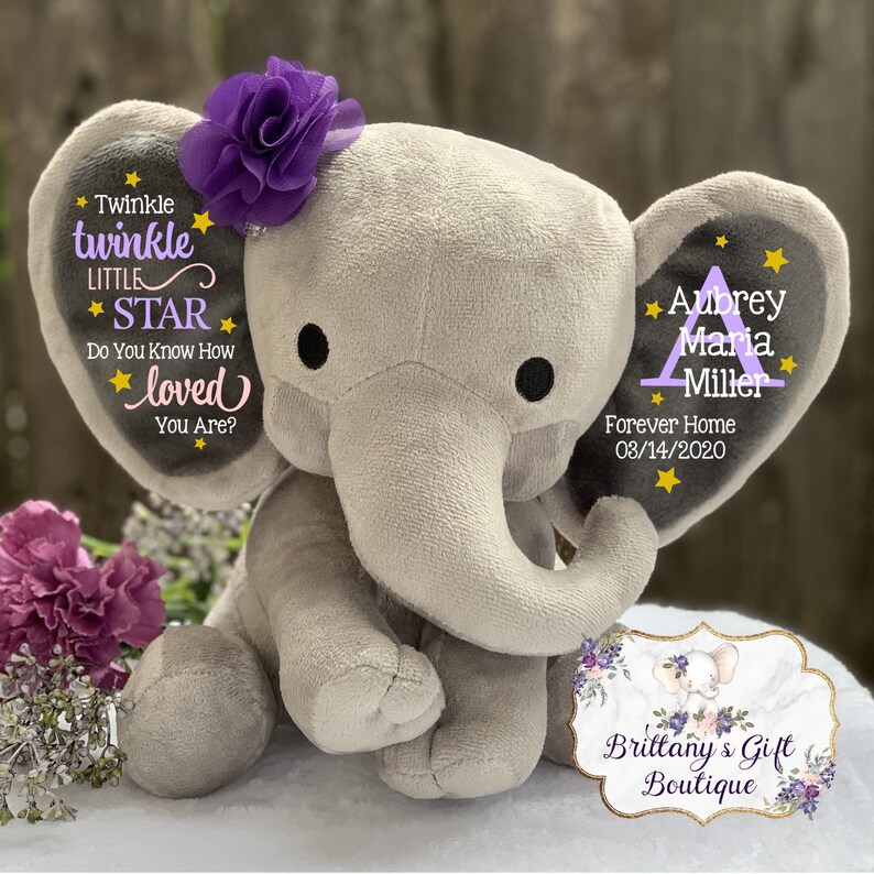 May include: A gray plush elephant toy with a purple fabric flower on its head. The left ear features the text Twinkle twinkle LITTLE STAR Do You Know How loved You Are? with yellow stars. The right ear displays the name Aubrey Maria Miller.