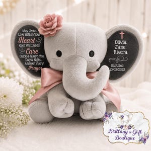 May include: A grey plush elephant toy featuring a pink satin ribbon and a pink flower on its head. The left ear displays the text: May Jesus Live Within Your Heart & Care Guide & Guard You, Day & Night, Answer Every Prayer. The right ear displays: Olivia Jane Rivera, a cross, and Baptized 10/18/2023.