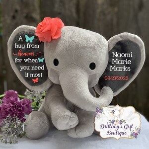 Puede incluir: Un elefante de peluche gris con una flor coral en la cabeza. La oreja del elefante tiene el texto "A hug from heaven for when you need it most." La otra oreja tiene el nombre "Naomi Marie Marks" y la fecha "03/27/2022".