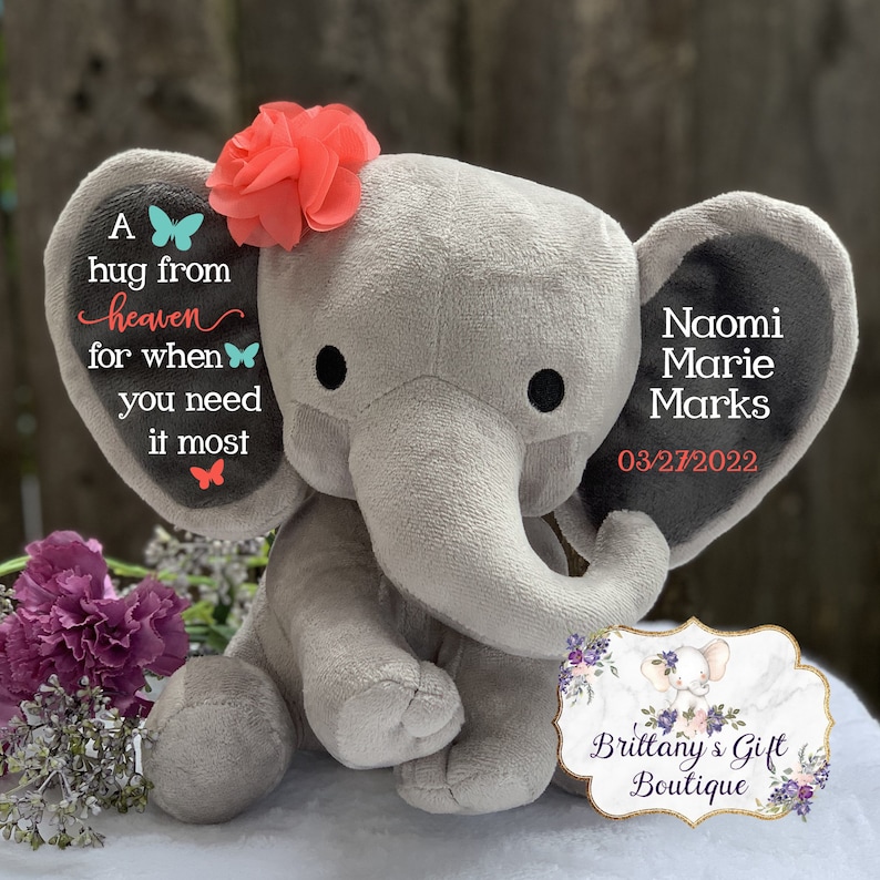 Puede incluir: Un elefante de peluche gris con una flor coral en la cabeza. La oreja del elefante tiene el texto "A hug from heaven for when you need it most." La otra oreja tiene el nombre "Naomi Marie Marks" y la fecha "03/27/2022".