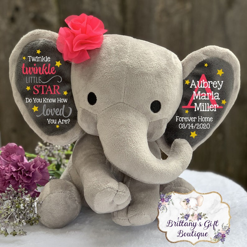 May include: A gray plush elephant toy with a pink fabric flower on its head. The left ear features the text Twinkle twinkle little star do you know how loved you are with yellow stars. The right ear displays the name Aubrey Maria Miller and date.