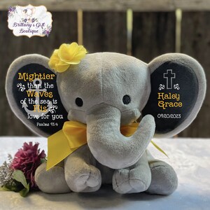 May include: A gray plush elephant toy featuring a yellow ribbon around its neck and a yellow flower on its head. The left ear displays an anchor icon and the text Mightier than the waves of the sea is His love for you Psalms 93:4. The right ear shows a cross icon, the name Haley Grace, and the date 09/20/2023.