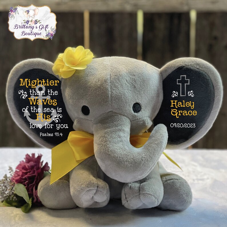 May include: A gray plush elephant toy featuring a yellow ribbon around its neck and a yellow flower on its head. The left ear displays an anchor icon and the text Mightier than the waves of the sea is His love for you Psalms 93:4. The right ear shows a cross icon, the name Haley Grace, and the date 09/20/2023.