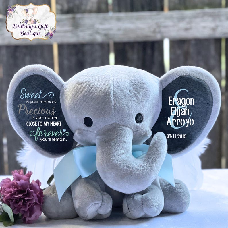 May include: A plush gray elephant with a light blue bow, featuring text on its ears. One ear reads "Sweet is your memory, Precious is your name, Close to my heart, forever you'll remain." The other ear has names and a date.