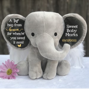 Puede incluir: Un elefante de peluche gris con orejas grandes, alas de plumas blancas y el texto "A hug from heaven for when you need it most" en una oreja. La otra oreja dice "Sweet Baby Marks 03/27/2022". Una margarita rosa est&aacute; delante.