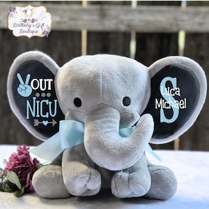 May include: A gray plush elephant toy with a blue ribbon. One ear has the text "OUT NICU" with a peace sign and arrow. The other ear has the letter "S" with the name "Luca Michael" underneath.