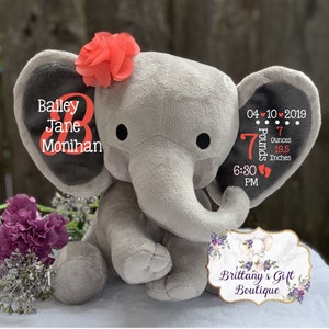 May include: A gray plush elephant toy featuring a coral fabric flower on its head. The left ear displays the name Bailey Jane Monihan in white and coral text. The right ear shows birth details: 04-10-2019, 7 Pounds 7 Ounces, 19.5 Inches, 6:30 PM.