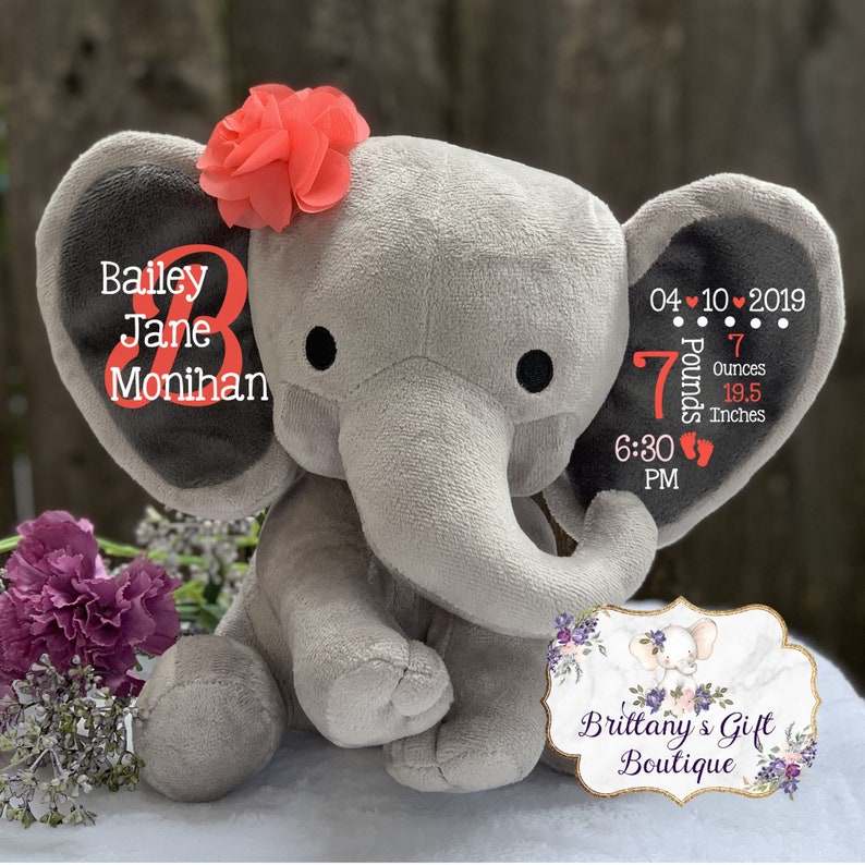 May include: A gray plush elephant toy featuring a coral fabric flower on its head. The left ear displays the name Bailey Jane Monihan in white and coral text. The right ear shows birth details: 04-10-2019, 7 Pounds 7 Ounces, 19.5 Inches, 6:30 PM.