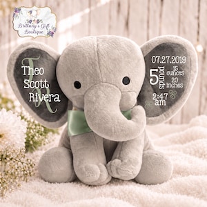 May include: A plush gray elephant toy with a light green satin bow. The left ear features the name Theo Scott Rivera in white text. The right ear displays birth details: 07.27.2019, 5 pounds 15 ounces, 20 inches, and 2:47 am in white text.