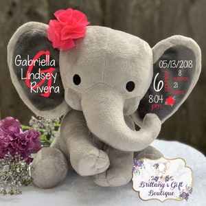 K&ouml;nnte beinhalten: Ein graues Pl&uuml;schelefant-Stofftier mit einer leuchtend rosa Stoffblume auf dem Kopf. Das linke Ohr zeigt den Namen Gabriella Lindsey Rivera in roter Schrift. Das rechte Ohr zeigt 05/13/2018, 6 pounds 8 ounces, 21 inches und 8:04 pm in Rot und Wei&szlig;.