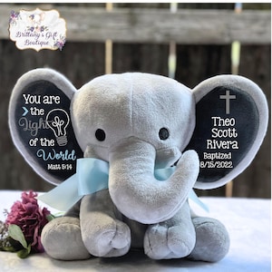 May include: A gray plush elephant with a light blue bow. The elephant's ears have text that reads "You are the Light of the World" and "Theo Scott Rivera Baptized 8/15/2022". A cross is on one ear. The elephant is a baptism gift.