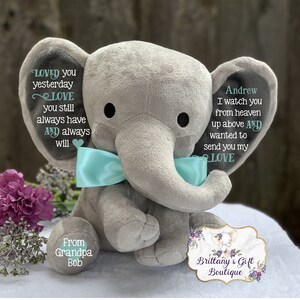 Puede incluir: Un elefante de peluche gris con un lazo azul claro. El elefante tiene un mensaje en cada oreja: "Loved you yesterday, Love you still, always have and always will" y "Andrew, I watch you from heaven up above and wanted to send you my love". El elefante est&aacute; sentado sobre una superficie blanca con una peque&ntilde;a etiqueta redonda que dice "From Grandpa Bob" y una etiqueta m&aacute;s grande que dice "Brittany's Gift Boutique".