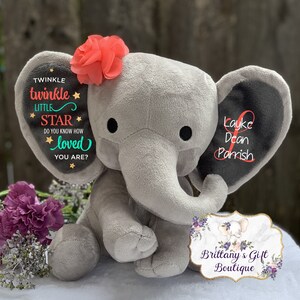 Puede incluir: Un elefante de peluche gris con una flor coral en la cabeza. Las orejas del elefante tienen el texto "Twinkle Twinkle Little Star" y un nombre. Un logotipo floral es visible.