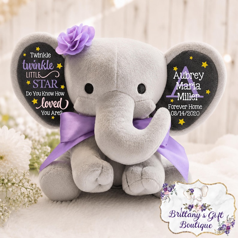 May include: A plush gray elephant toy featuring a purple fabric flower on its head and a purple satin ribbon around its neck. The ears are printed with yellow stars and text reading Twinkle twinkle little star and Aubrey Maria Miller.