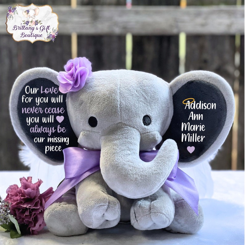 Może przedstawiać: Szary pluszowy słoń z fioletowymi skrzydłami i fioletową kokardką. Słoń ma czarne filcowe ucho z tekstem "Our Love for you will never cease you will always be our missing piece" i drugie czarne filcowe ucho z tekstem "Addison Ann Marie Miller".