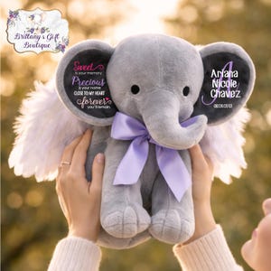 May include: A gray plush elephant with a purple bow, white feathered wings, and text on its ears. One ear reads "Sweet is your memory, Precious is your name, Close to my heart forever you'll remain." The other ear reads "Ariana Nicole Chavez 08/24/2013."