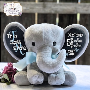 May include: A gray plush elephant with black eyes and a light blue ribbon. The elephant's ears have embroidered text: "Theo Scott Rivera" and birth details: 5 pounds, 15 ounces, 20 inches, 2:47 am, and the date 07.27.2019.