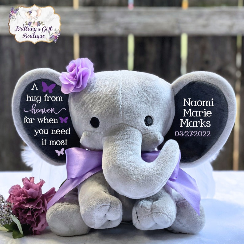 Puede incluir: Un elefante de peluche gris con un lazo y una flor morados. El elefante tiene texto en las orejas, incluyendo "Naomi Marie Marks 03/27/2022" y "A hug from heaven for when you need it most."
