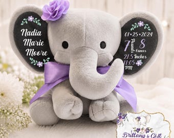May include: A plush gray elephant toy featuring a purple ribbon bow and a purple flower on its head. The ears are personalized with white text including the name Nadia Marie Moore, birth date 01-25-2024, 7 pounds 8 ounces, 21.5 inches, and 11:05 PM.