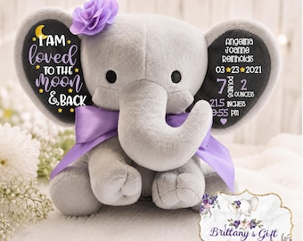 May include: A grey plush elephant toy with a purple satin ribbon and a purple fabric flower on its head. The ears feature printed text: I AM loved TO THE MOON & BACK, and birth details including 7 pounds 2 ounces and 21.5 inches.