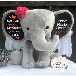 Puede incluir: Un elefante de peluche gris con una flor rosa y alas de plumas blancas. El elefante tiene texto en sus orejas que dice "A hug from heaven for when you need it most" y "Naomi Marie Marks 03/27/2022". Un logotipo de boutique de regalos est&aacute; en la parte inferior.