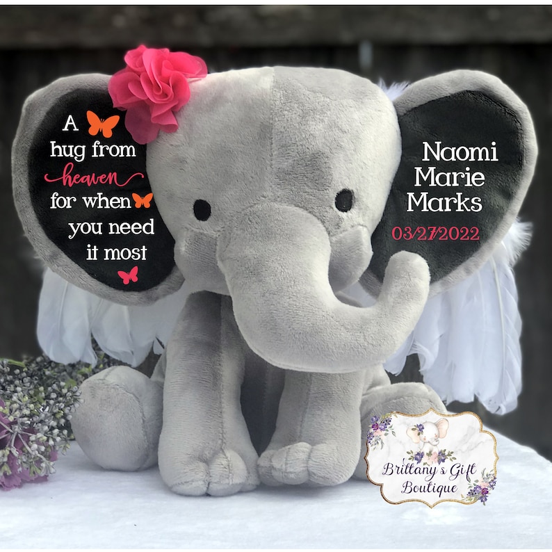 Puede incluir: Un elefante de peluche gris con una flor rosa y alas de plumas blancas. El elefante tiene texto en sus orejas que dice "A hug from heaven for when you need it most" y "Naomi Marie Marks 03/27/2022". Un logotipo de boutique de regalos est&aacute; en la parte inferior.