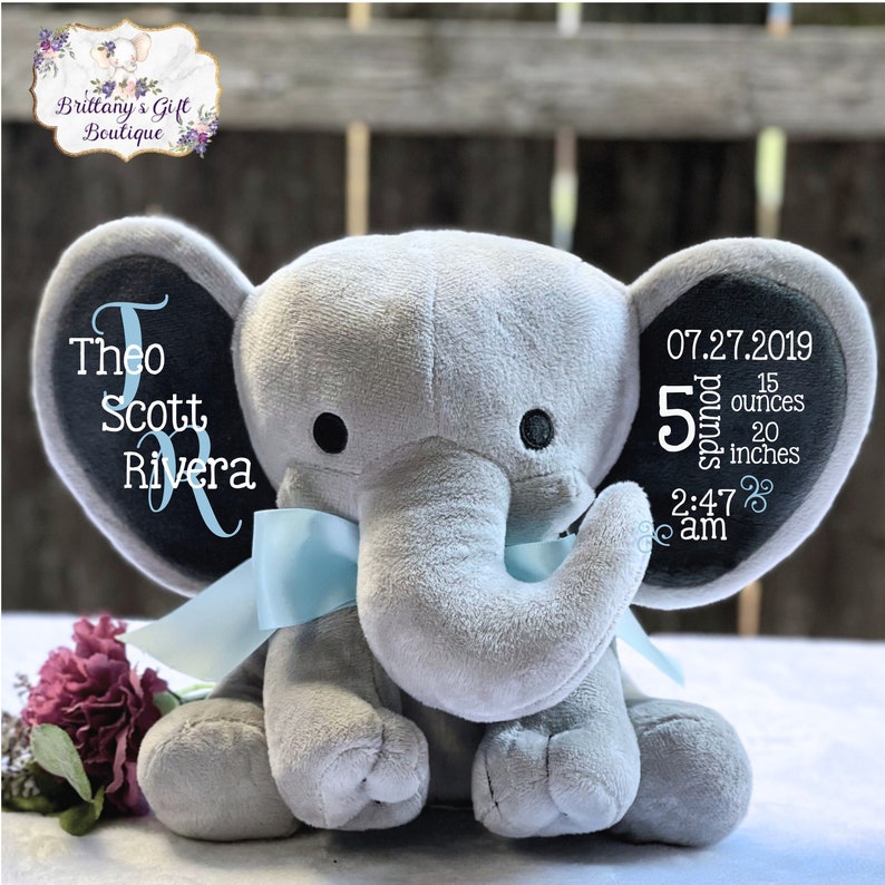 Puede incluir: Un elefante de peluche gris con ojos negros y una cinta azul claro. Las orejas del elefante tienen texto bordado: "Theo Scott Rivera" y detalles de nacimiento: 2,27 kg, 50,8 cm, 2:47 am, y la fecha 27.07.2019.