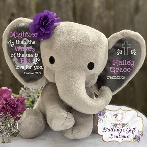 May include: A plush gray elephant toy featuring a purple fabric flower on its head. The left ear displays an anchor graphic with the text Mightier than the waves of the sea is His love for you Psalms 93:4. The right ear shows a cross, the name Hailey Grace, and the date 09/20/2023.