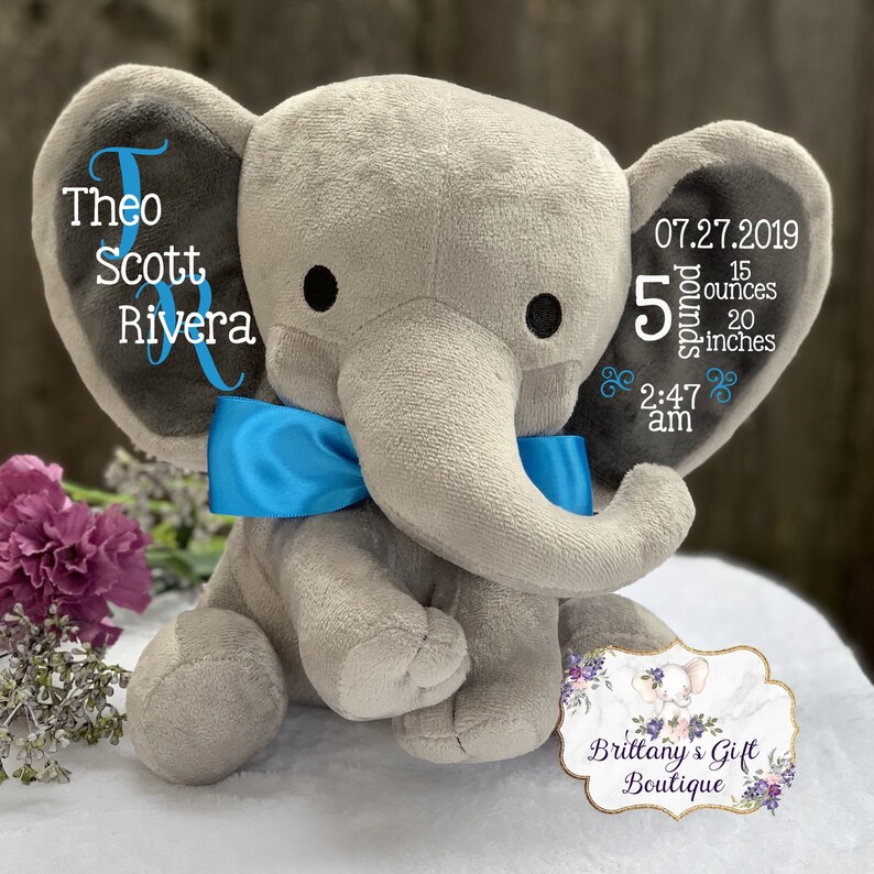 Puede incluir: Un peluche de elefante gris con una pajarita azul. Las orejas del elefante muestran texto bordado con el nombre "Theo Scott Rivera" y detalles de nacimiento: 07.27.2019, 2,27 kg, 50,8 cm y 2:47 am. Una etiqueta de regalo es visible.