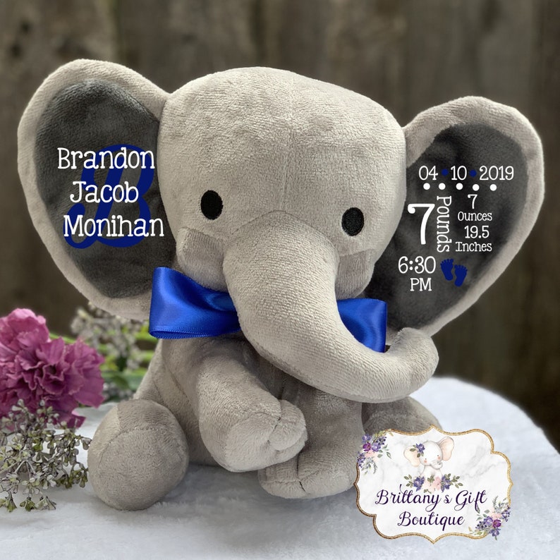 May include: A gray plush elephant toy featuring a blue satin bow around its neck. The left ear displays the name Brandon Jacob Monihan in blue and white text. The right ear shows birth details: 04 10 2019, 7 Pounds 7 Ounces, 19.5 Inches, and 6:30 PM.