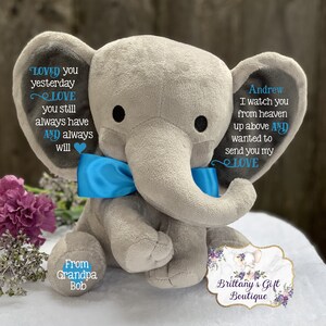 Puede incluir: Un elefante de peluche gris con una pajarita azul. El elefante tiene texto bordado que dice "Loved you yesterday, Love you still, always have and always will." y "Andrew, I watch you from heaven up above and wanted to send you my love." El elefante est&aacute; sentado sobre una superficie blanca con una etiqueta que dice "From Grandpa Bob" y "Brittany's Gift Boutique".