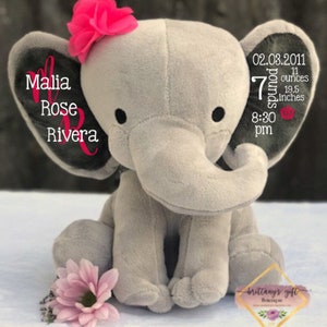 May include: A plush gray elephant toy with a pink flower on its head and a pink flower at its feet. The elephant's ear displays the name "Malia Rose Rivera" in pink script. The other ear shows the birth details: 02.03.2011, 7 pounds, 11 ounces, 19.5 inches, and 8:30 pm.