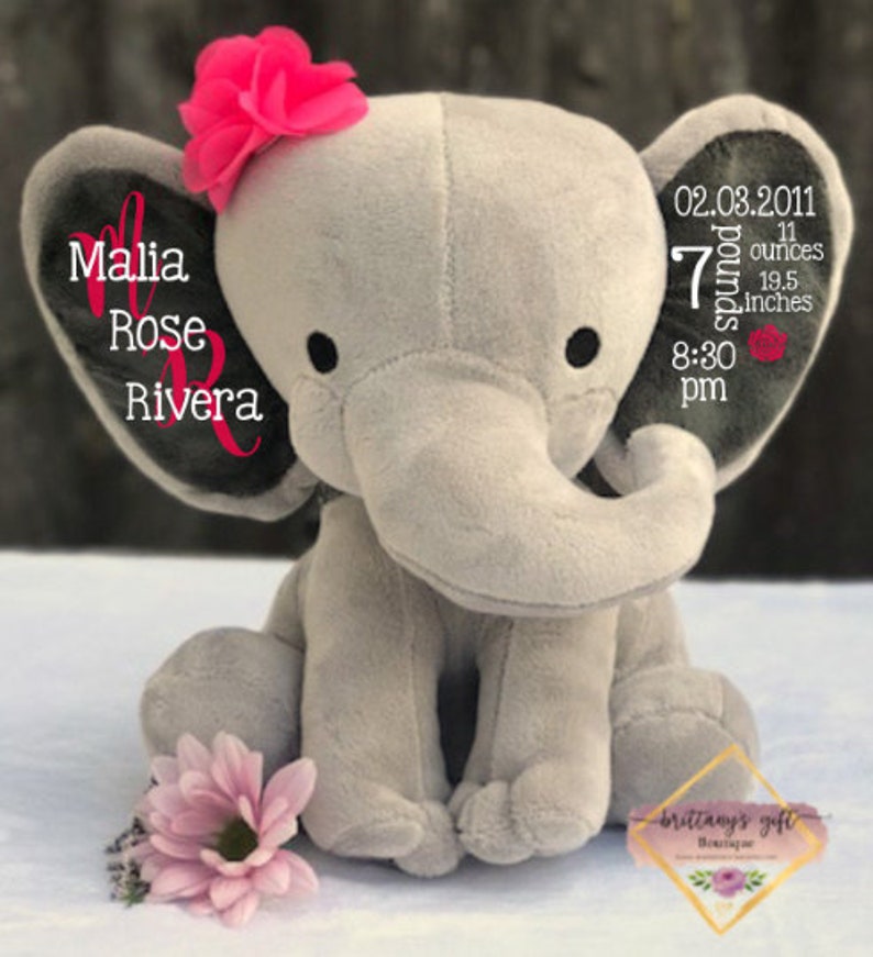 May include: A plush gray elephant toy with a pink flower on its head and a pink flower at its feet. The elephant's ear displays the name "Malia Rose Rivera" in pink script. The other ear shows the birth details: 02.03.2011, 7 pounds, 11 ounces, 19.5 inches, and 8:30 pm.