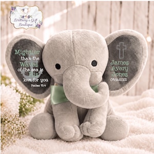 May include: A plush gray elephant toy with a light green satin bow. The left ear features an anchor graphic and the text: Mightier than the Waves of the sea is His love for you Psalms 93:4. The right ear displays a cross and the name James Avery Jones 09/16/2023.