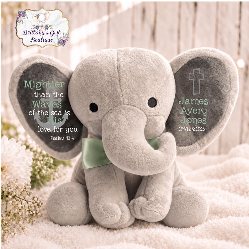 May include: A plush gray elephant toy with a light green satin bow. The left ear features an anchor graphic and the text: Mightier than the Waves of the sea is His love for you Psalms 93:4. The right ear displays a cross and the name James Avery Jones 09/16/2023.