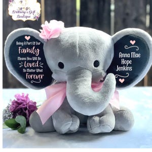 May include: A gray plush elephant with pink ears and a pink bow. The elephant has a black heart on one ear with the text "Being A Part Of Our Family Means You Will Be Loved No Matter What Forever". The other ear has a black heart with the text "Anna Mae Hope Jenkins".