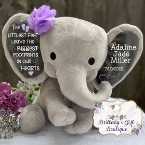 Op de afbeelding: Een grijze pluche olifant met paarse bloemenaccenten. De oren van de olifant zijn geborduurd met de tekst "The littlest feet leave the biggest footprints in our hearts" en "Adaline Jade Miller 7/10/2022". Een bord met de tekst "Brittany's Gift Boutique" staat op de voorgrond.