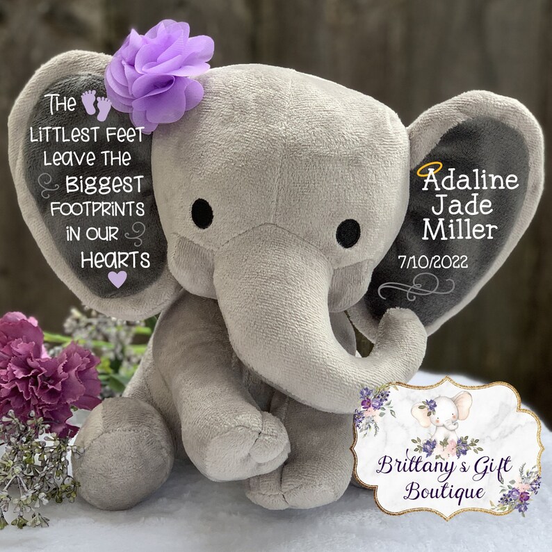 Op de afbeelding: Een grijze pluche olifant met paarse bloemenaccenten. De oren van de olifant zijn geborduurd met de tekst "The littlest feet leave the biggest footprints in our hearts" en "Adaline Jade Miller 7/10/2022". Een bord met de tekst "Brittany's Gift Boutique" staat op de voorgrond.