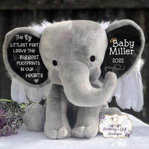 Op de afbeelding: Een grijze pluche olifant met witte vleugels en zwarte oren. Het ene oor heeft een zwart hart met de tekst "The littlest feet leave the biggest footprints in our hearts". Het andere oor heeft de tekst "Baby Miller 2022" in zwart met een gouden halo boven de naam.