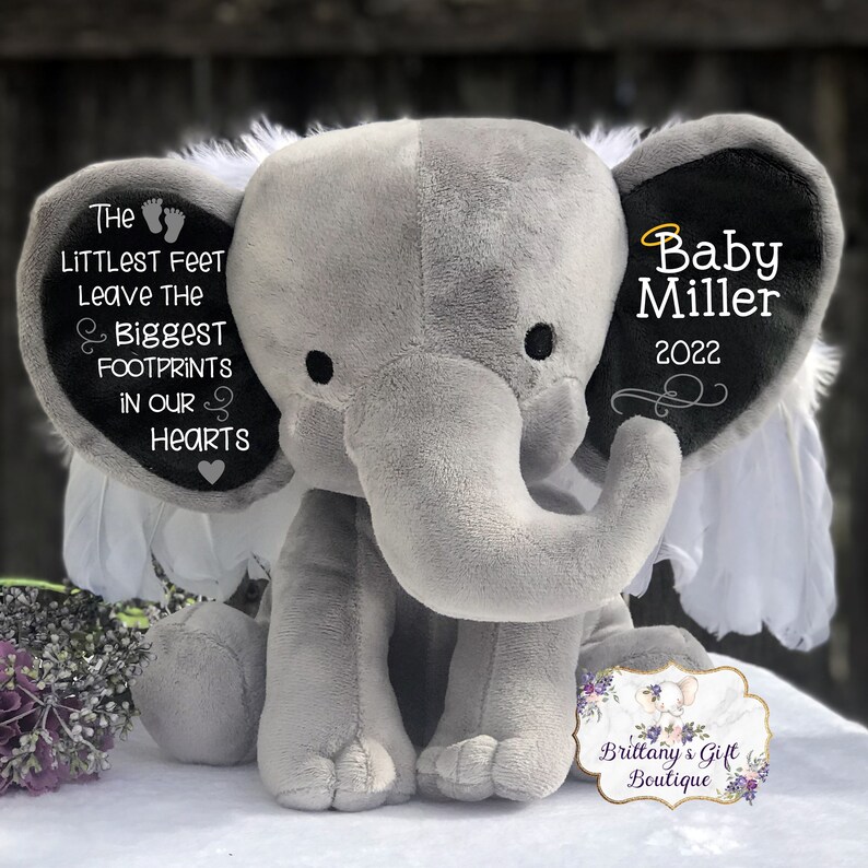 Op de afbeelding: Een grijze pluche olifant met witte vleugels en zwarte oren. Het ene oor heeft een zwart hart met de tekst "The littlest feet leave the biggest footprints in our hearts". Het andere oor heeft de tekst "Baby Miller 2022" in zwart met een gouden halo boven de naam.