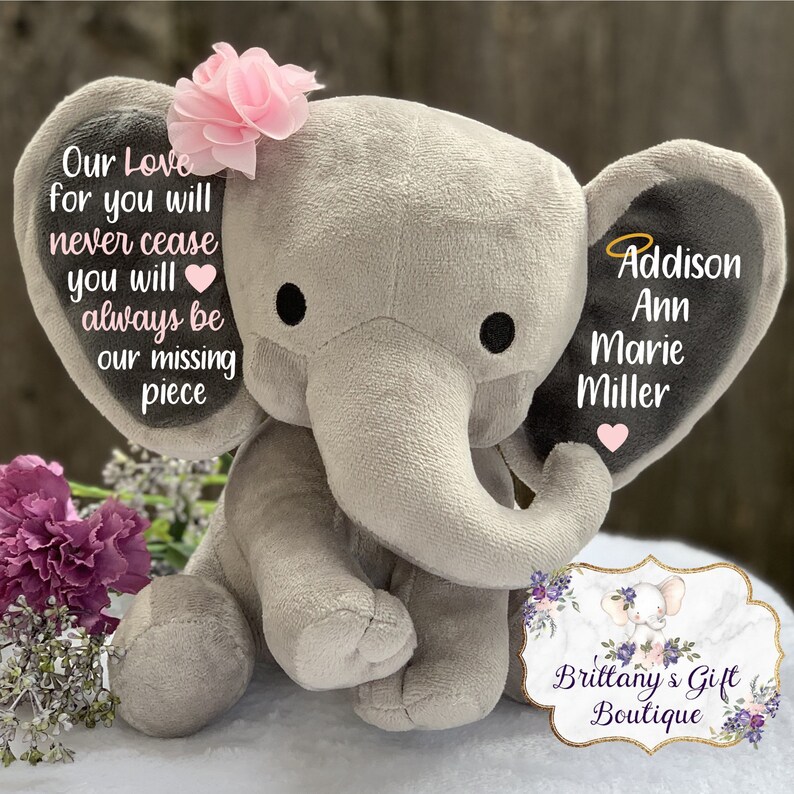 Może przedstawiać: Szary pluszowy słoń z r&oacute;żowym kwiatem na głowie. Uszy słonia mają czarne tło z białym tekstem, kt&oacute;ry brzmi "Our Love for you will never cease you will always be our missing piece" na jednym uchu i "Addison Ann Marie Miller" na drugim uchu. Słoń siedzi na białym tle z tabliczką z napisem "Brittany's Gift Boutique" przed nim.