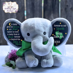 Puede incluir: Un elefante de peluche gris con alas verdes y un lazo verde. Las orejas del elefante est&aacute;n decoradas con c&iacute;rculos de fieltro negro con estrellas blancas y texto. La oreja izquierda dice "A baby gone to Heaven ★ An Angel taken to soon A baby that is Loved forever ★ Beyond the Stars & Moon". La oreja derecha dice "Forever in our ★ Hearts Baby Jackson 2023 ★".