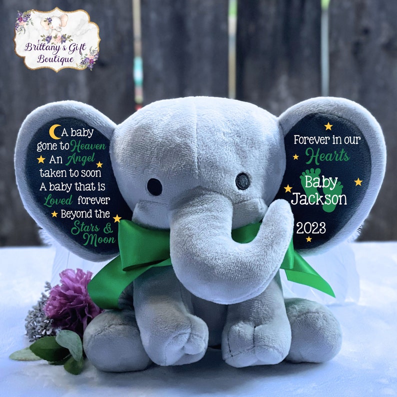 Puede incluir: Un elefante de peluche gris con alas verdes y un lazo verde. Las orejas del elefante est&aacute;n decoradas con c&iacute;rculos de fieltro negro con estrellas blancas y texto. La oreja izquierda dice "A baby gone to Heaven ★ An Angel taken to soon A baby that is Loved forever ★ Beyond the Stars & Moon". La oreja derecha dice "Forever in our ★ Hearts Baby Jackson 2023 ★".